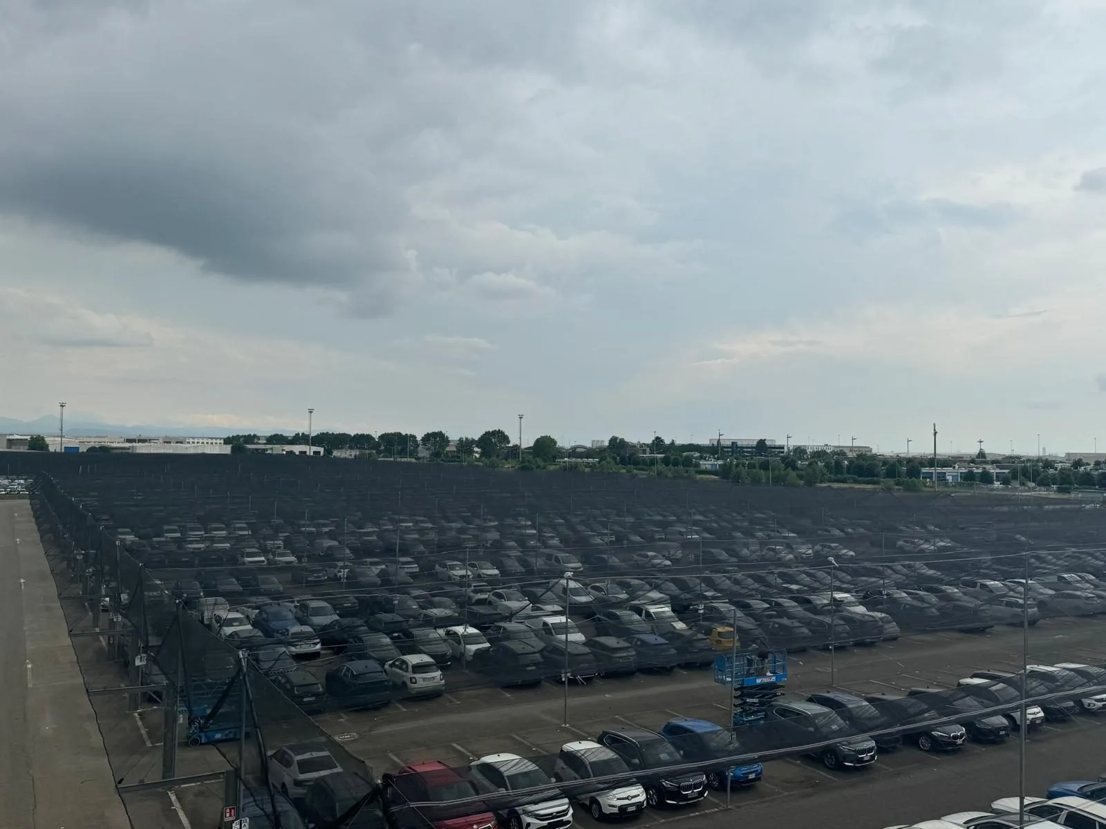 Grand parking de concessionnaire avec des centaines de véhicules neufs protégés par un filet anti-grêle noir tendu sur structure métallique, sous un ciel nuageux